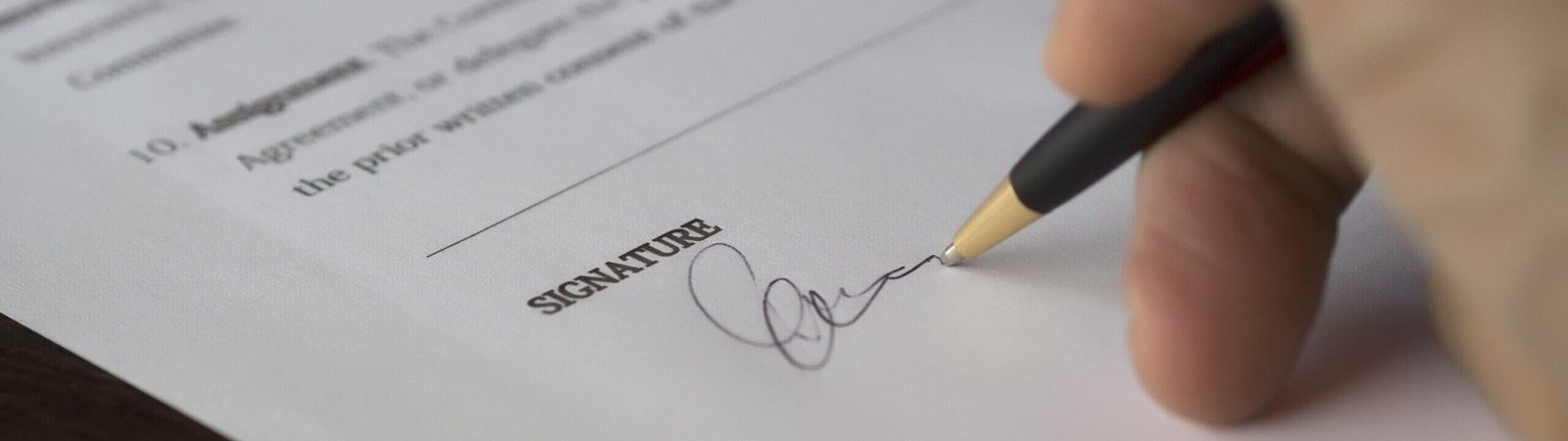 A hand signs a formal contract with a pen on a wooden desk.